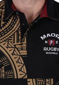 T-shirt de rugby noir avec des motifs tribaux dorés, un col intérieur rouge et un logo brodé blanc "MAORI NZ RUGBY RUCKFIELD." Présente des coutures contrastées.