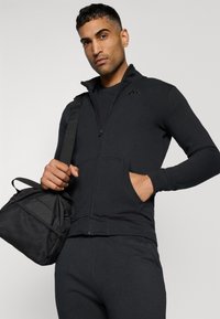 Jaqueta atlética preta com zíper e bolsos, usada com uma camisa preta correspondente. O modelo segura uma mala de ginásio texturizada preta com alças.