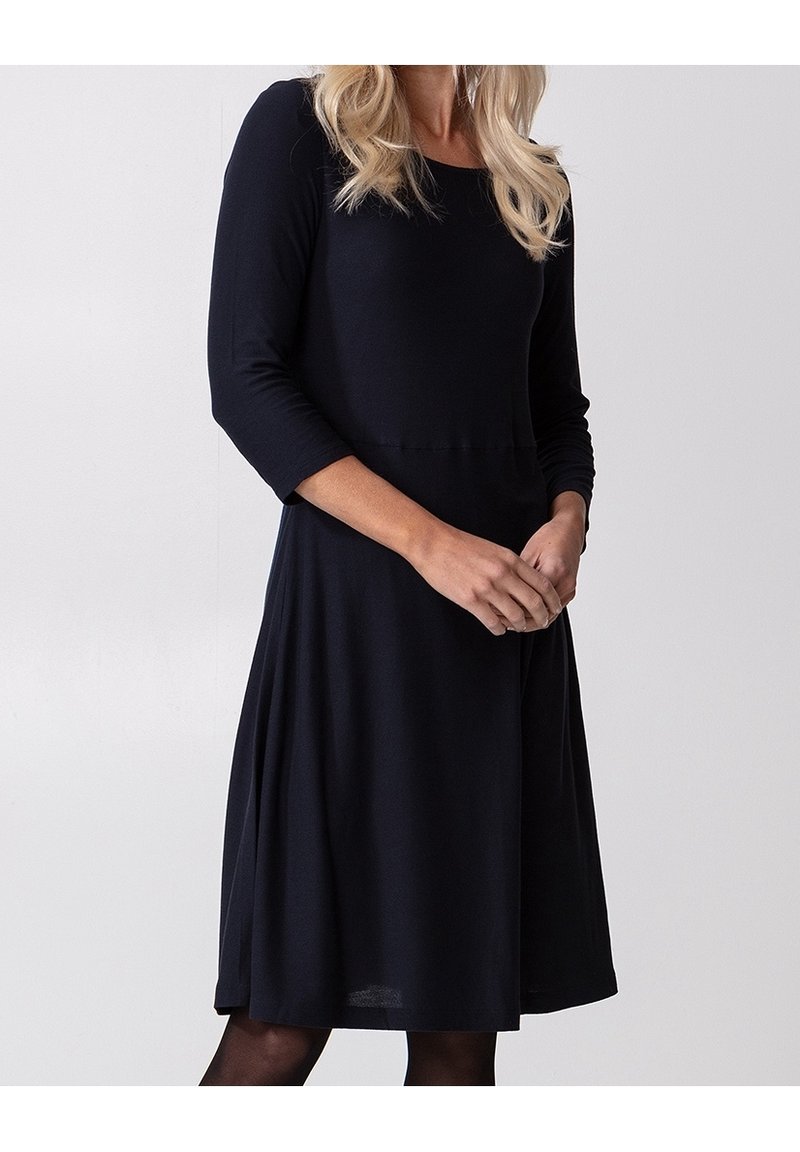 Femme portant une robe noire mi-longue avec des manches trois quarts, debout, les mains jointes lâchement devant un fond uni.