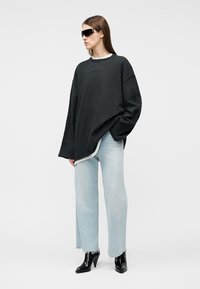 Frau trägt einen übergroßen schwarzen Pullover, hellblaue Jeans mit weitem Bein, schwarze Stiefeletten mit Absatz und große schwarze Sonnenbrille, steht mit einer Hand in der Tasche.