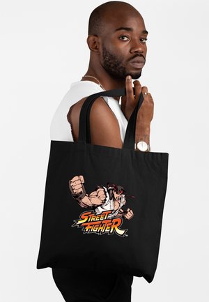 Man die een wit mouwloos shirt draagt, houdt een zwarte tas met een Street Fighter-personage en logo ontwerp vast.