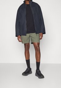 Veste zippée bleu marine foncé sur un t-shirt noir, associée à un short vert olive, des chaussettes noires et des chaussures de sport noires. Design simple, coupe décontractée.