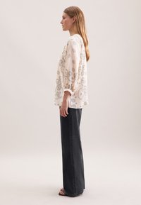 Lys beige bluse med blomstermønster, puffede ærmer og en afslappet pasform, parret med mørkegrå vide bukser. Glat tekstur, afslappet design.
