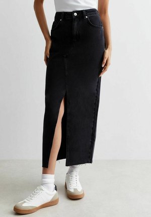 Jupe midi en denim noir avec une fente devant, ourlet effiloché, portée avec des baskets blanches et des chaussettes, montrée de la taille aux pieds.
