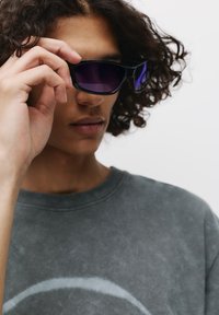 Czarne okulary przeciwsłoneczne z purpurowymi soczewkami, kątowy design i podwyższone zauszniki, trzymane w dłoni przeciwko szarym bluzie tie-dye.