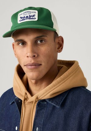 Młody mężczyzna noszący zielono-białą czapkę Levi Strauss, beżową bluzę z kapturem oraz niebieską kurtkę jeansową, patrzy prosto w obiektyw z neutralnym wyrazem twarzy.