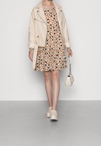 Kremowa kurtka z sztucznej skóry nałożona na sukienkę w geometryczny wzór w odcieniach beżu i czerni, z falbaną na dole, uzupełniona jasnymi, wsuwanymi butami oraz małą torebką.