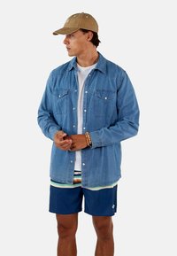 Hellblaues Denim-Hemd mit zwei Brusttaschen, Knopfverschluss und langen Ärmeln, getragen über einem weißen T-Shirt und gestreiften Shorts.
