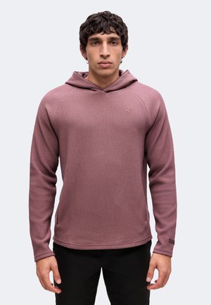 Young man wearing a textured mauve hooded long-sleeve shirt and black pants, standing against a plain light background.