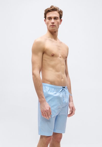 Νεαρός άνδρας με σγουρά μαλλιά, ντυμένος με ανοιχτό μπλε και λευκό ριγέ μαγιό, στέκεται μπροστά σε μια απλή ανοιχτόχρωμη επιφάνεια.