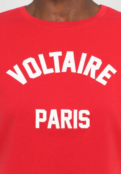 Červené bavlněné tričko s výrazným bílým nápisem, který v oblouku hlásá "VOLTAIRE" nad "PARIS," kulatým výstřihem a hladkou texturou.