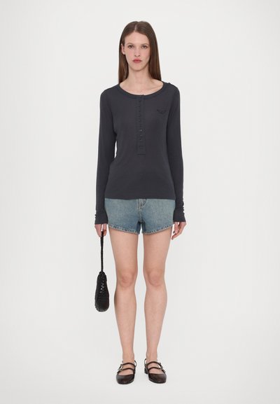 Langärmliges, dunkelblaues Oberteil mit Knopfdetails, kombiniert mit hellen Jeansshorts. Schwarze strukturierte Tasche in der Hand gehalten; braune Schuhe sind zu sehen.