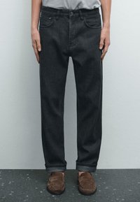 Jeans in denim grigio scuro con vestibilità ampia, cinque tasche e fondo risvoltato, abbinati a mocassini marroni su una superficie testurizzata.