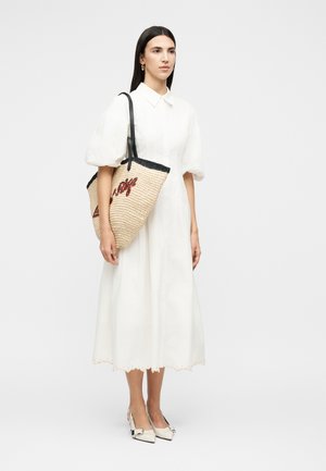 Žena v bielych midi šatách s nafúknutými rukávmi nesúca pletenú slamennú tašku s čiernym lemom a červeným nápisom, obutá v bielych podpätkoch so sponami.