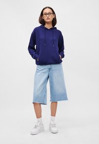 Námornícka mikina s viazacím šnúrkami a prednou vreckom, v kombinácii so svetlomodrými džínovými šortkami po kolená a bielymi teniskami, s uvoľneným strihom.