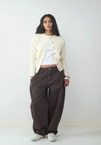 Cremefarbener Strickcardigan mit Rundhalsausschnitt, braune weite Hose und weiße Sneaker. Nennenswerte Merkmale sind goldene Knöpfe und kontrastierende Nähte.