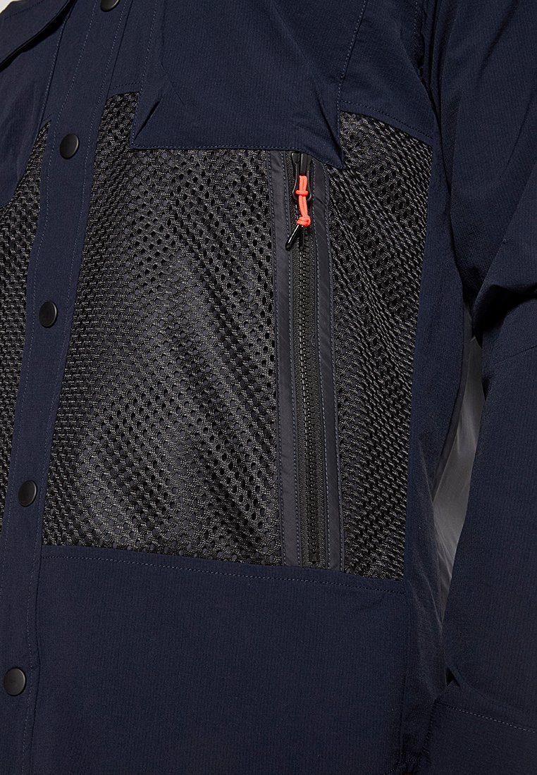 Marinejacke mit einem strukturierten schwarzen Netzpaneel, ausgestattet mit einem Reißverschluss und einem orangen Zugband sowie schwarzen Knopfakzenten auf der Vorderseite.