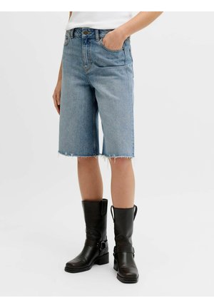 Personne portant un short en jean bleu clair longueur genou avec des bords bruts et des bottes en cuir noires mi-mollet, debout sur un fond blanc.