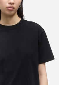 Νεαρός με μακριά σκούρα μαλλιά φορώντας μια απλή μαύρη κοντομάνικη μπλούζα με στρογγυλή λαιμόκοψη και ένα ασημένιο κρίκο στο αυτί σε ανοιχτό γκρι φόντο.