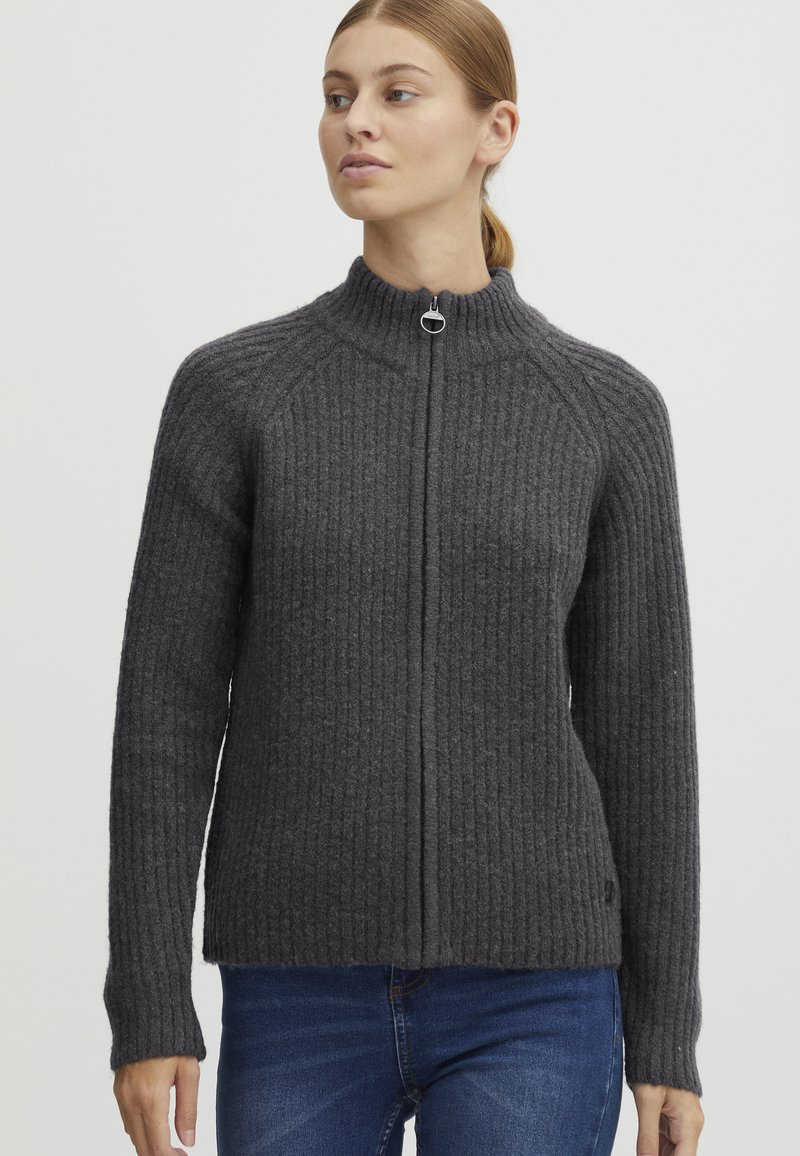Grå ribbestrikket genser med høy krage og frontglidelås. Har lange ermer og en litt løs passform. Stoffet ser teksturert og varmt ut.