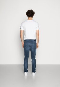 Camiseta blanca de mangas cortas con rayas negras en las mangas, combinada con jeans ajustados azules y zapatillas blancas, vista desde la parte de atrás.