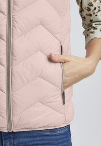 Gilet matelassé rose clair avec un motif en chevrons diagonal, fermeture éclair devant et poche latérale. Matériau doux, léger éclat et col élégant.