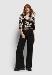 Schwarze Bluse mit Blumenmuster und Kragen, kombiniert mit hochgeschnittenen, weiten schwarzen Hosen. Brauer Gürtel mit großer silberner Schnalle und schwarzen Plateaustiefeln.