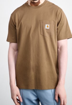 Mand iført en brun Carhartt pocket t-shirt og lyseblå bukser, der står foran en ensfarvet hvid baggrund.