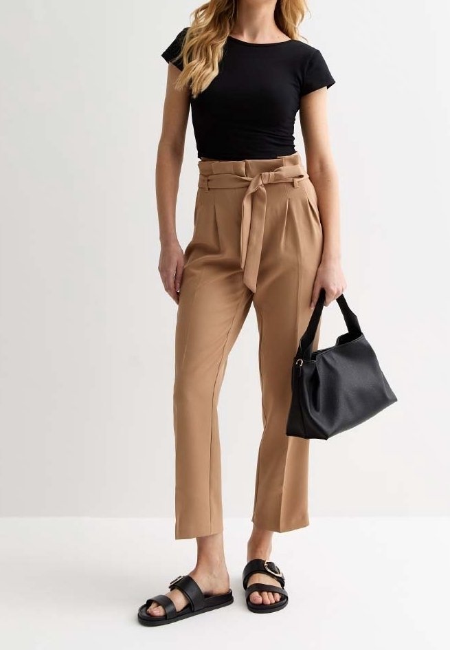 Zwarte, nauwsluitende T-shirt, hoge beige broek met een strikceintuur, zwarte handtas en zwarte sandalen met een gespdetail, staande op een neutrale achtergrond.