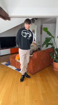 Νέος άνδρας με μαύρο φουτερ με τυπώματα, μπεζ παντελόνι και μαύρα μποτάκια στέκεται στο σαλόνι με καναπέ σε σκουρόχρωμη σκουριά και μεγάλο φυτεμένο φυτό.