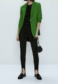 Zielona marynarka na czarnej bluzce, zestawiona z czarnymi legginsami i loafersami. Marynarka ma wyraźnie zarysowane ramiona i jeden guzik.