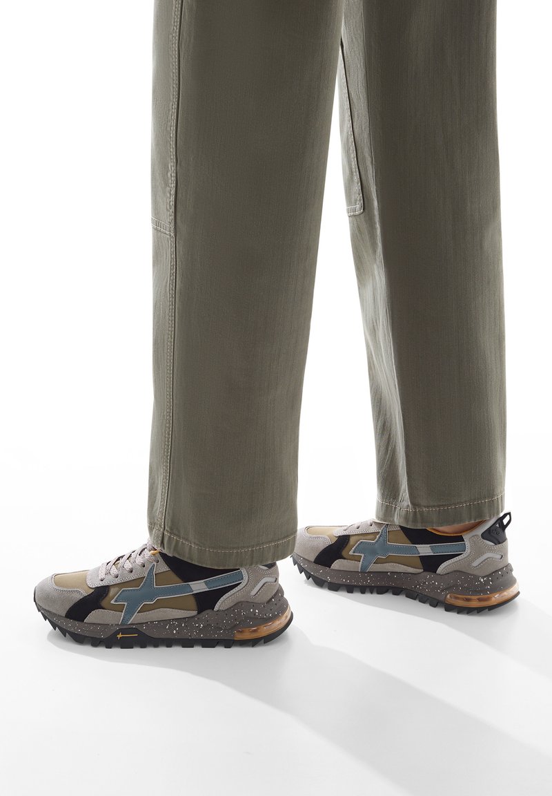 Sneakers con una combinazione di suede grigio, nero e marrone chiaro, con un accento blu, suola testurizzata e lacci visibili. Abbinati a pantaloni verde oliva.
