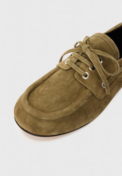 Zapato de barco de ante en verde oliva con ojales plateados y cordones a juego. Presenta una puntera redondeada y un cuello acolchado para mayor comodidad.