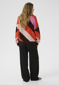 Pull en tricot multicolore avec des rayures diagonales rouge, rose, marron et crème. Coupe ample avec un col rond, associé à un pantalon large noir.