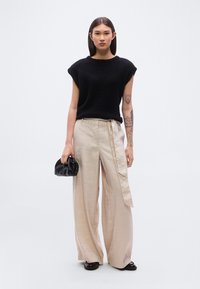 Μαύρο πλεκτό πουλόβερ με κοντά μανίκια, σε συνδυασμό με μπεζ παντελόνι από λινό με φαρδιά πόδια και πλαϊνή πινακίδα. Κρατείται μια μικρή μαύρη τσάντα.