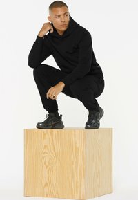 Black hooded sweatshirt and joggers, both made of soft fabric. Model wearing black sneakers, squatting on a wooden cube, with a smooth texture.