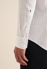 Camicia bianca a maniche lunghe con motivo a quadri blu, dotata di polsini bottonati e dettaglio ricamato di rosa sulla manica. Tessuto morbido.