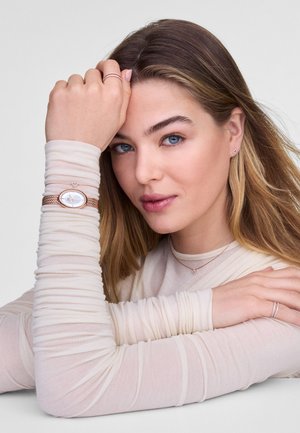 Montre en or rose avec un cadran ovale sur un bracelet en maille, assortie à une bague en argent et des boucles d'oreilles en forme de cœur. Le modèle porte un haut léger à manches longues texturé.