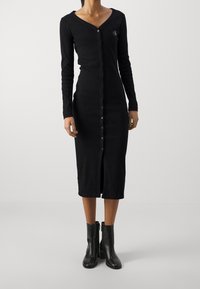 Musta pitkähihaine mekko, jossa on V-pääntie, nappi etuosa ja halkio helmaosassa. Valmistettu pehmeästä kankaasta, jossa on hienovarainen logotieto rinnassa.