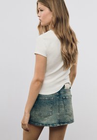Junge Frau mit langen, lockigen Haaren, die ein weißes T-Shirt mit kurzen Ärmeln und einen blauen Denim-Minirock trägt, steht vor einem schlichten Hintergrund.