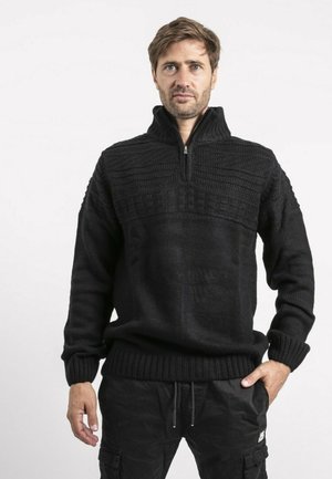 Homme portant un pull en maille texturée noire avec un col à demi-fermeture éclair et un pantalon cargo noir, debout avec une main dans la poche.