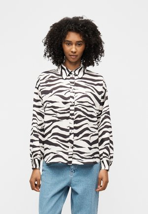 Femme aux cheveux bouclés portant une chemise boutonnée à imprimé zèbre noir et blanc et un jean bleu clair, debout devant un fond uni.