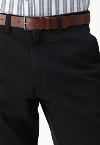 Pantalon noir en coton à la texture lisse, maintenu par une ceinture en cuir marron avec une boucle en métal argenté. Chemise à rayures horizontales visible.