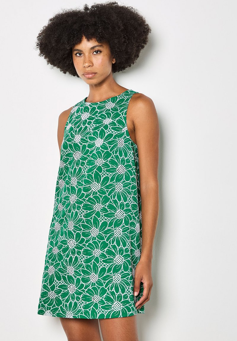 Femme aux cheveux bouclés portant une robe verte sans manches avec un motif floral blanc, debout devant un fond clair uni.