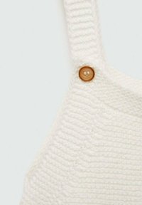 Valkoinen neulevaate, jossa on ribbikuvio ja pyöreä ruskea nappidetaili hihnassa. Lähikuva kankaasta ja kiinnityksestä.