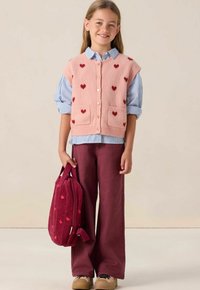 Un gilet en tricot rose avec des motifs de cœurs rouges, superposé sur une chemise à rayures bleues. Un pantalon large bordeaux et un sac à dos en velours côtelé assorti.