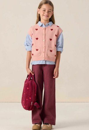 Roze gebreide vest met rode hartpatronen, gelaagd over een blauw gestreept shirt. Kastanjebruine wijde broek en een bijpassende corduroy rugzak.