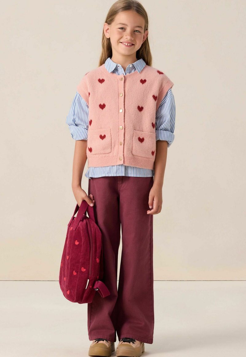 Un gilet en tricot rose avec des motifs de cœurs rouges, superposé sur une chemise à rayures bleues. Un pantalon large bordeaux et un sac à dos en velours côtelé assorti.