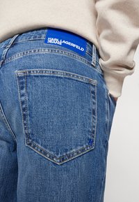 Sinine denimist teksapüksid sisaldavad silmapaistvat tagataskut, millel on kontrastne õmblus ja sinine nahast silt, millel on kirjas "KARL LAGERFELD JEANS."
