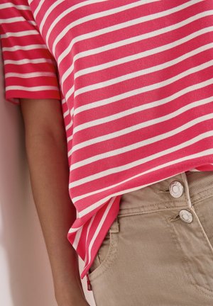 Henkilö, jolla on päällään punavalkoraidallinen lyhythihainen paita, joka on hieman puettu beigeihin nappihousuihin, arm suojaa vaaleaa seinää vasten.